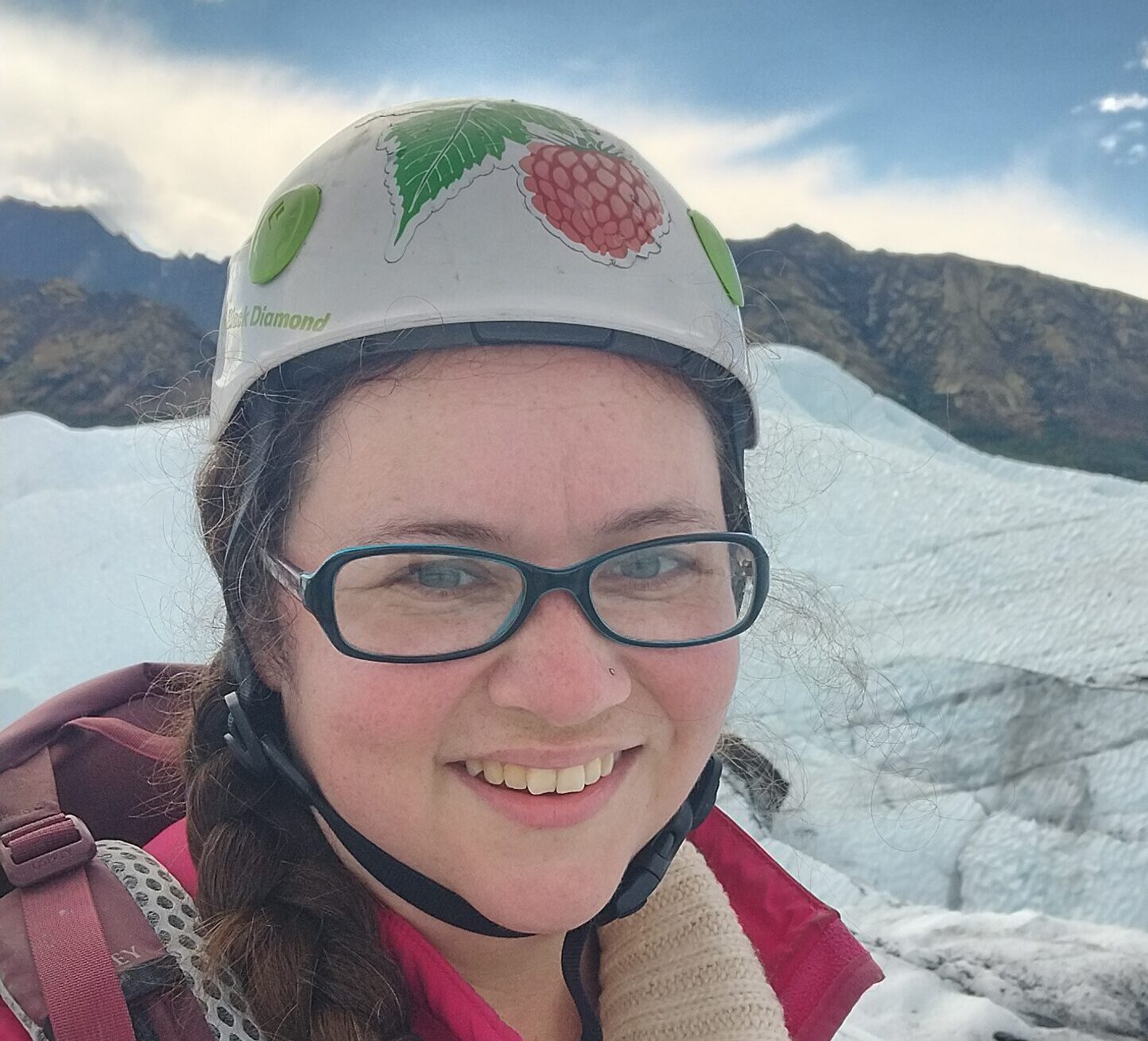 Michelle has long brown hair in a thick braid over her right shoulder. She has glasses, and is wearing a pink Salmon Berry Tours vest and a white helmet, with glacier ice in the background.