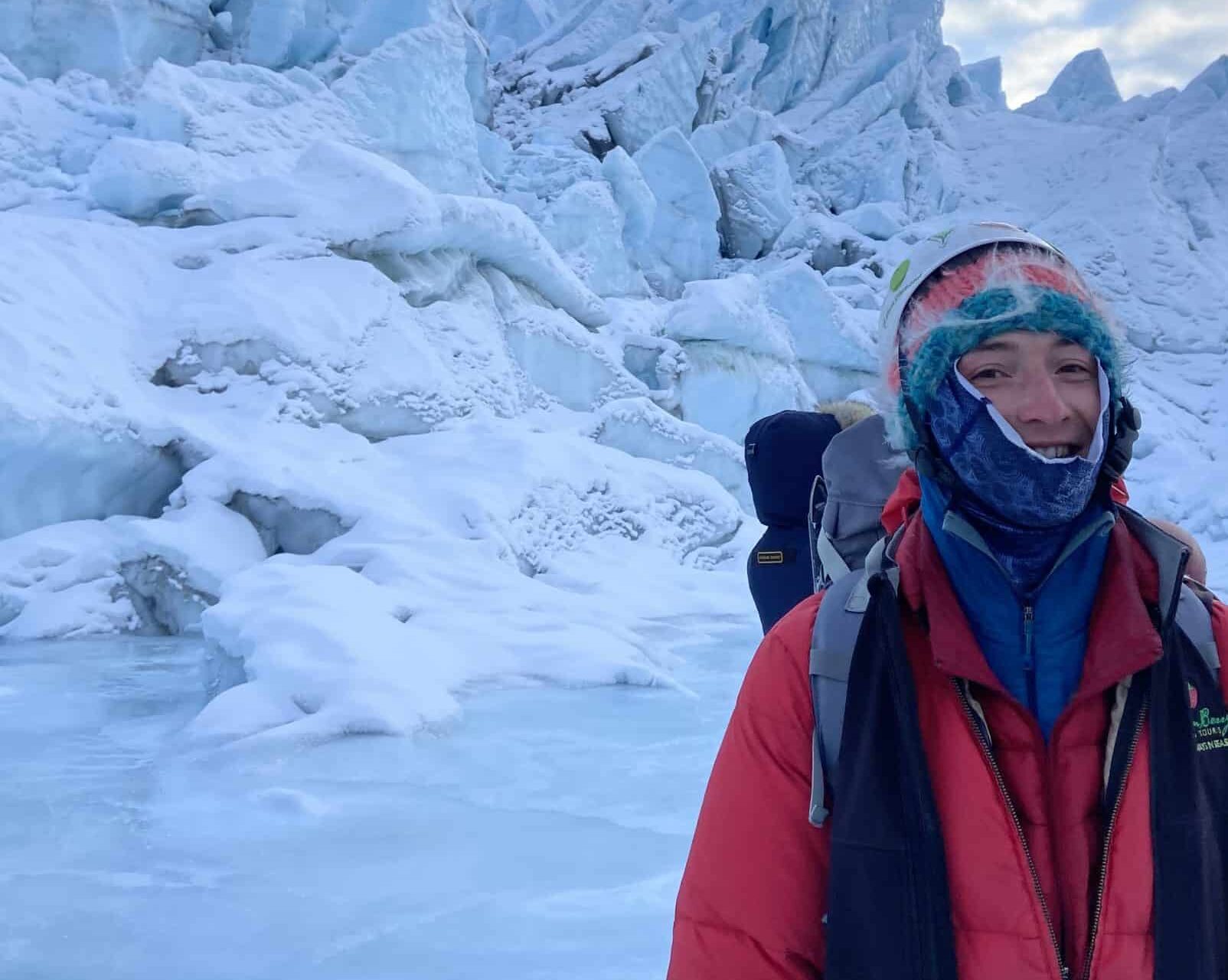 Josh looks very cold on front of a fully ice background, wearing an orange puffer parka, a black vest, a backpack with climbing gear showing, a blue face gater, and a very colorful hat.