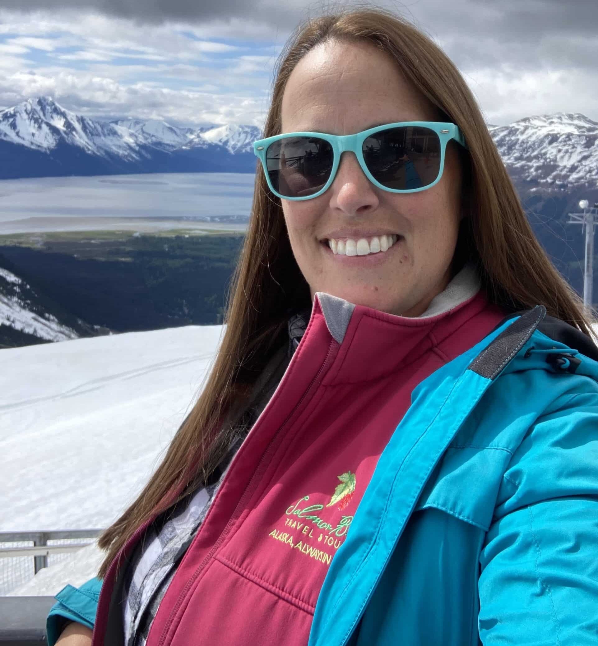 Jeri has long straight brown hair and is pictured in front of the view of Turnagain Arm from the top of Mt Alyeska. She is wearing a pink Salmon Berry Tours vest, with a blue jacket and matching blue sunglasses.