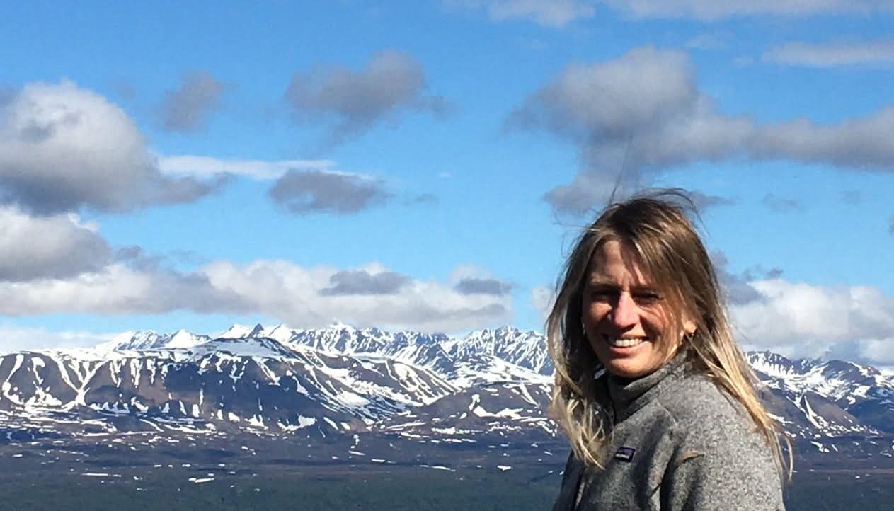 Patty has sandy blond hair and is pictured in a gray sweater with a stretch of the Alaska Range in the background