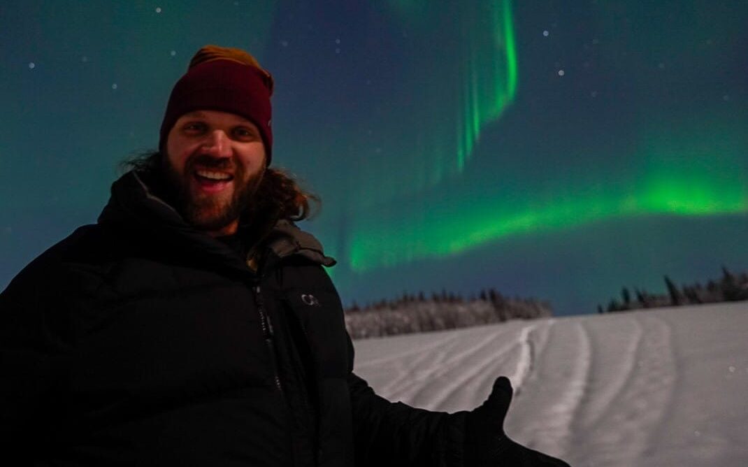 Harrison has medium length brown hair and is pictured at night, wearing a black jacket, a burgundy beanie hat, and he is standing on snow, smiling, with a green band of Aurora behind him.