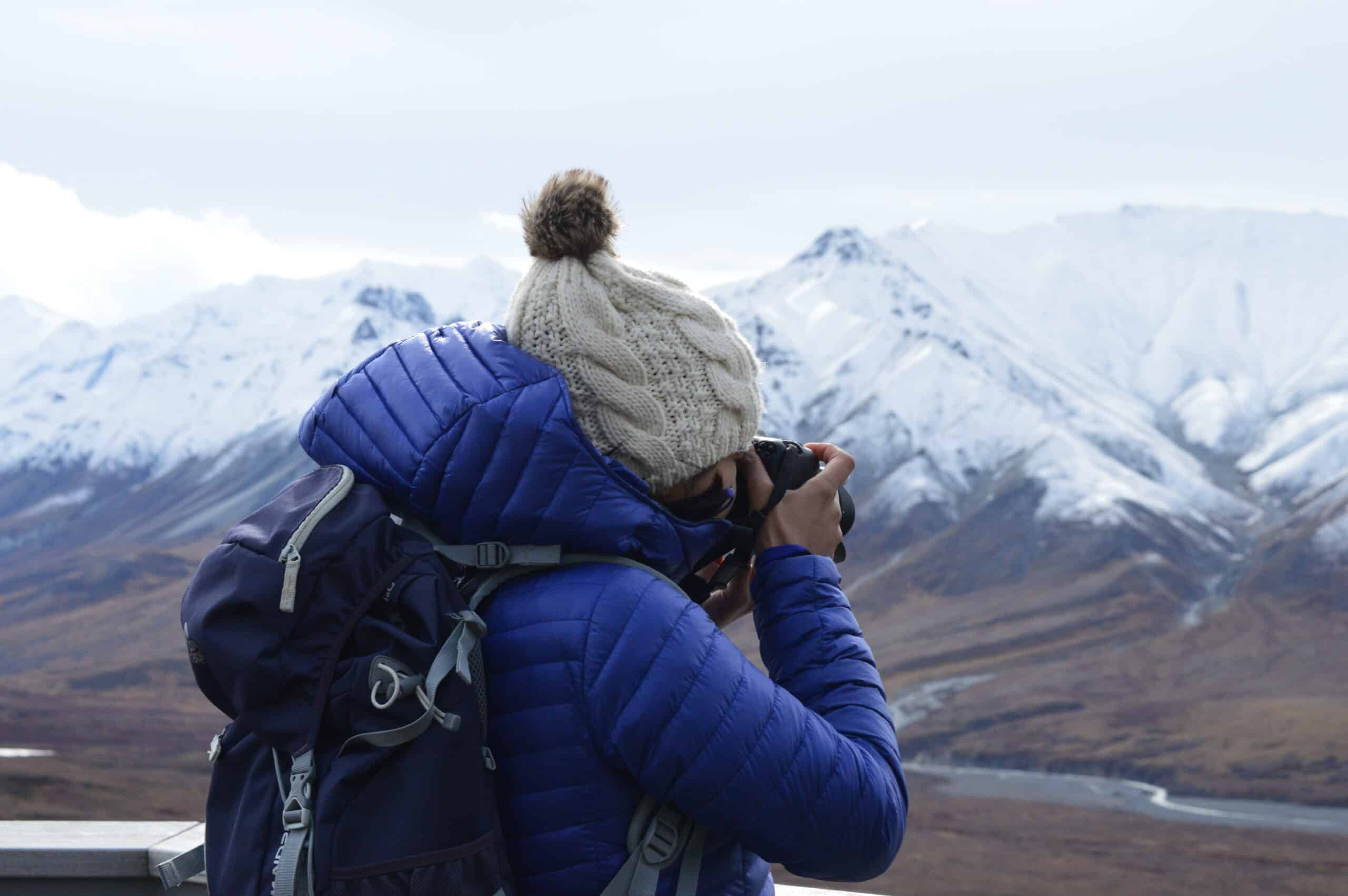 Photographer capturing a mountain landscape, wearing a blue jacket and wool hat.