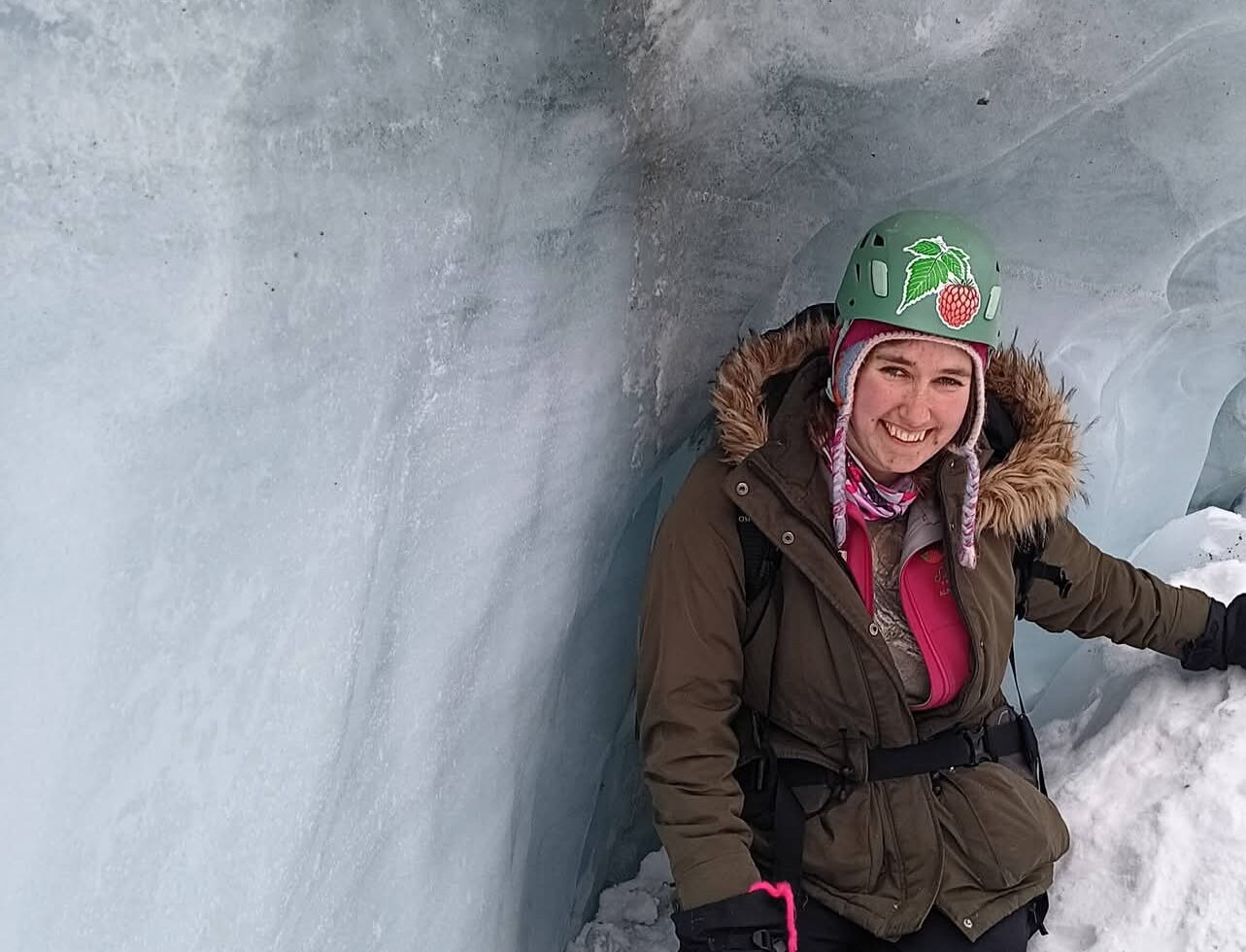 Kayla is smiling and wearing a parka, with a green helmet, crouching next to an ice wall on Matanuska Glacier