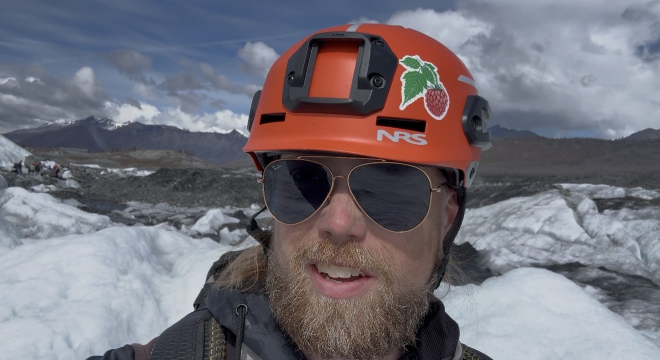Glacier Guide Derek is pictured with ice formations in the background, wearing an orange helmet with a beard and sunglasses