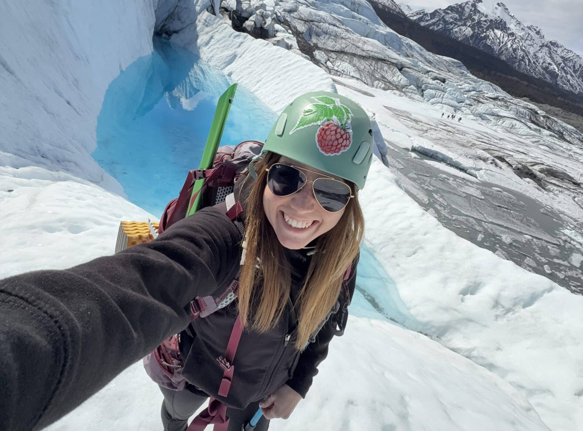 Glacier Guide Rachel has long brown hair, is wearing a green helmet and a backpack with climbing gear, and is standing on glacier ice in front of a blue meltwater pool, wearing her cool sunglasses and smiling